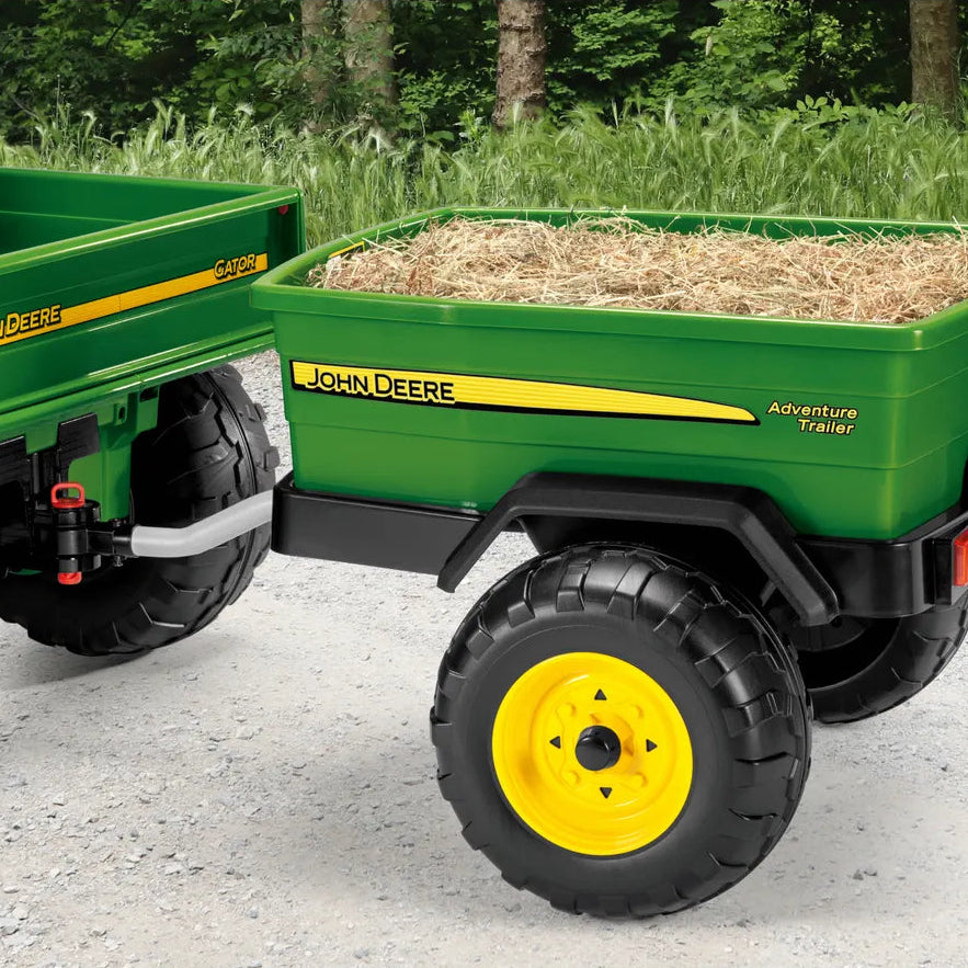 A green John Deere Adventure Trailer toy wagon with yellow wheels, shown attached to a matching green toy trailer on a gravel path with grass and trees in the background.